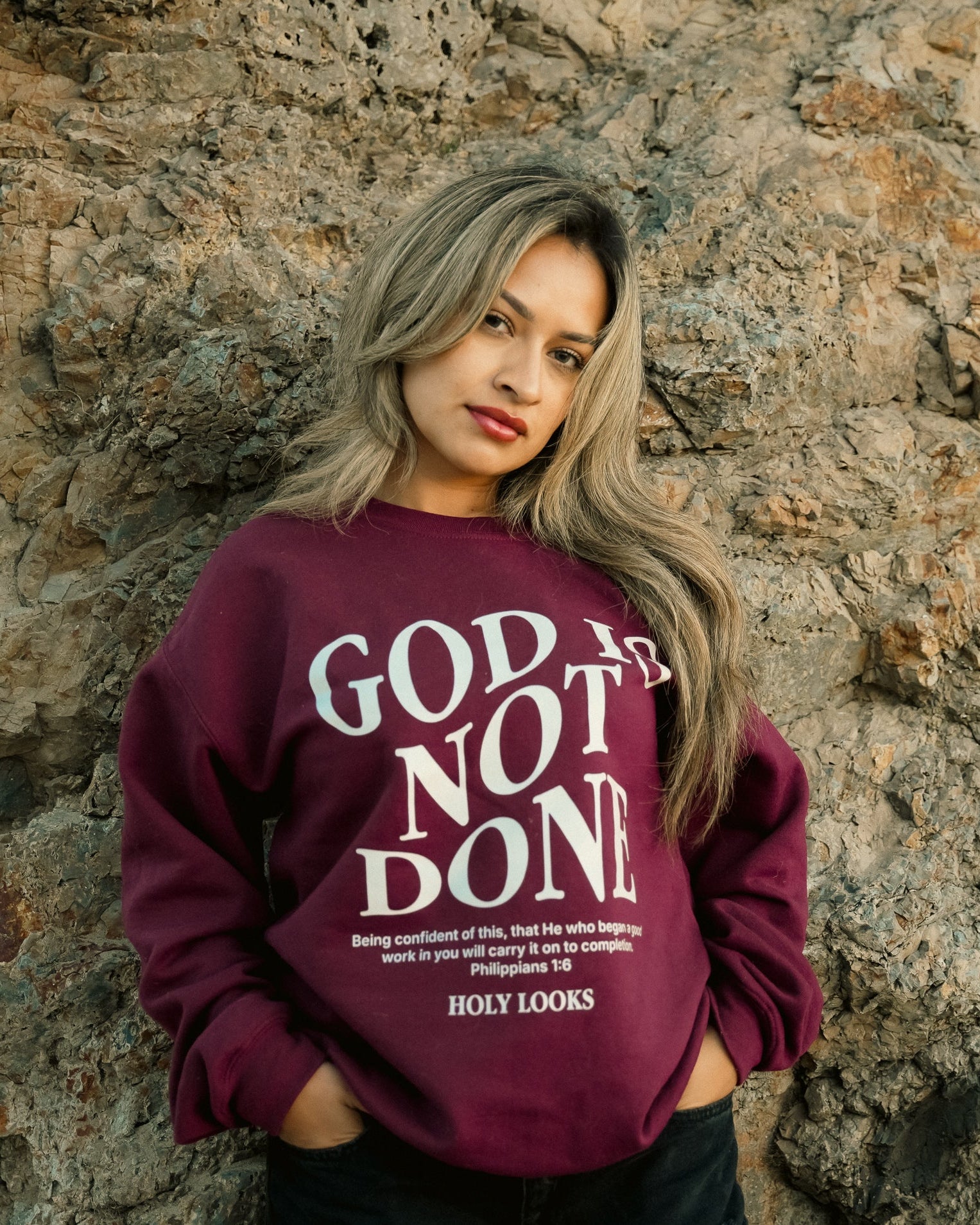 A woman with blonde-highlighted hair stands against a textured stone wall wearing a maroon crewneck sweatshirt with the words 'GOD IS NOT DONE' in white text, paired with black pants.