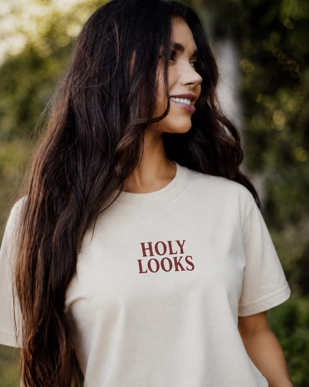 A woman with long dark hair wearing a cream-colored t-shirt with the words 'HOLY LOOKS' printed in dark red text on the chest, set against a blurred outdoor background of green foliage.