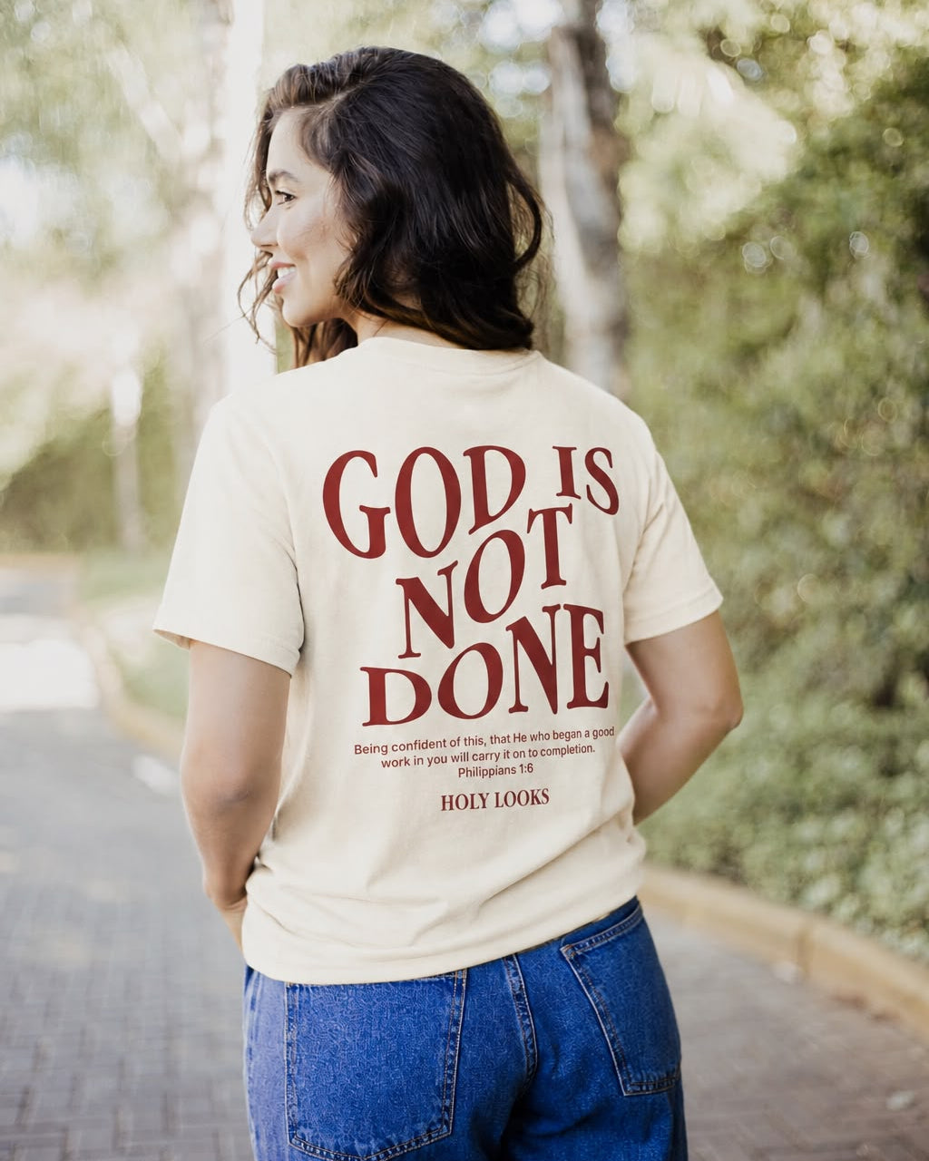 A woman wearing a cream-colored t-shirt with the text 'GOD IS NOT DONE' printed in dark red on the back, standing outdoors on a paved path with trees in the background.