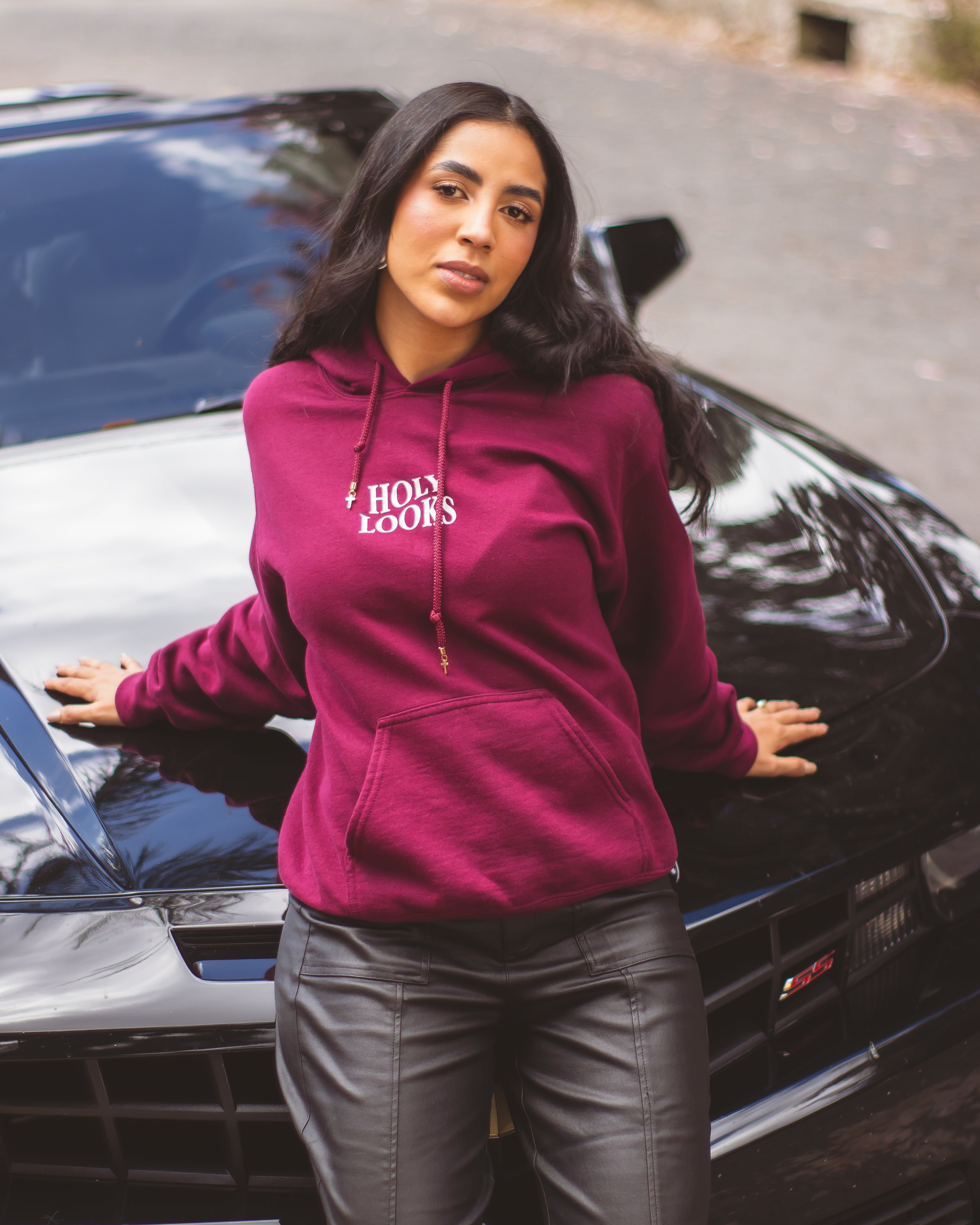 A woman wearing a magenta hoodie with 'HOLY LOOKS' printed in white on the chest, posing with a black car outdoors.