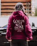 A person viewed from behind wearing a maroon hoodie with 'GOD IS NOT DONE' printed in white on the back, standing outdoors in front of a black car and wooden wall.