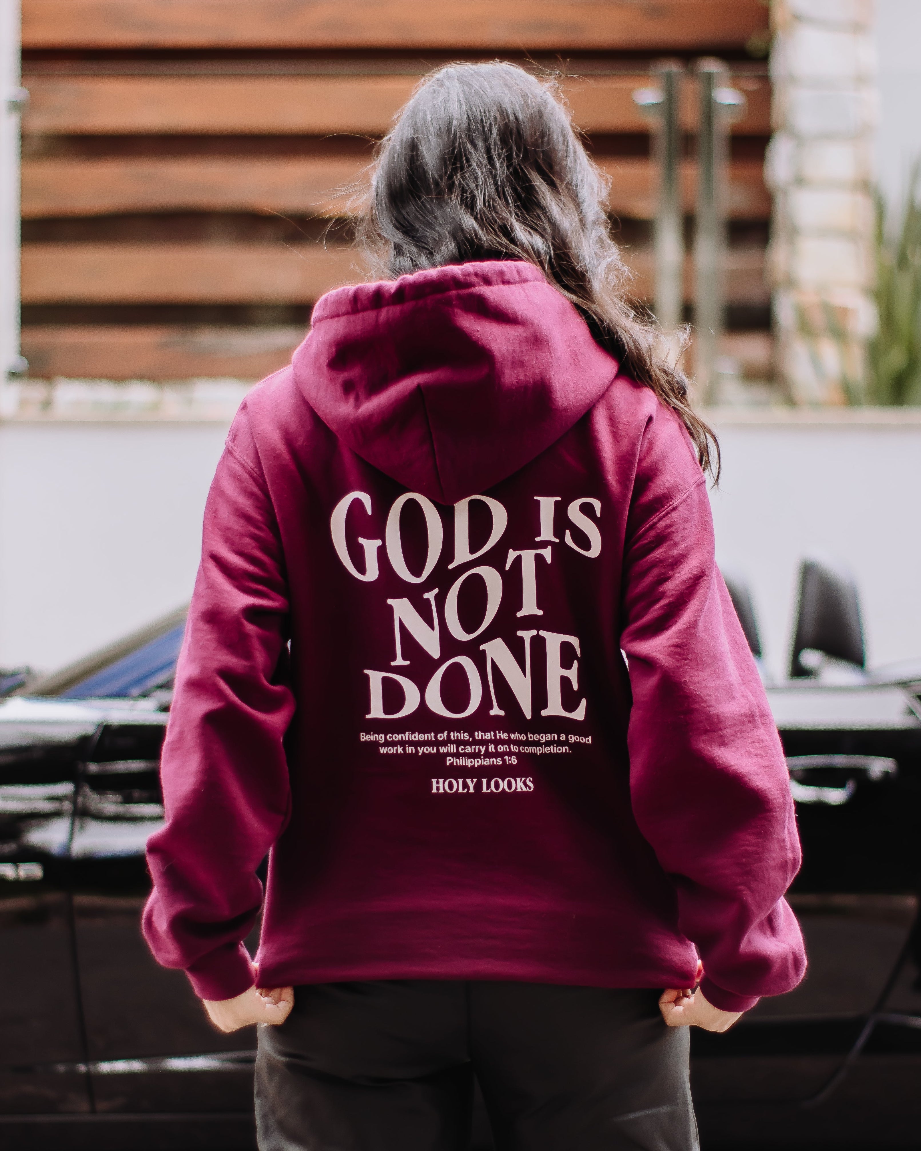 A person viewed from behind wearing a maroon hoodie with 'GOD IS NOT DONE' printed in white on the back, standing outdoors in front of a black car and wooden wall.