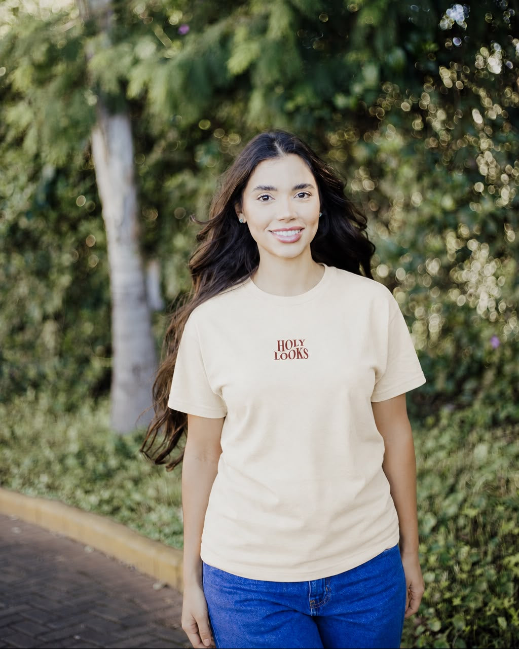 A woman with long dark hair stands outdoors on a path wearing a light beige crewneck t-shirt with 'BOY TOYS' printed in red on the chest, paired with blue jeans. The background is a soft-focus green garden.