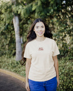 A woman with long dark hair stands outdoors on a path wearing a light beige crewneck t-shirt with 'BOY TOYS' printed in red on the chest, paired with blue jeans. The background is a soft-focus green garden.