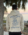A man viewed from behind wearing a cream-colored hoodie with a blue graphic print on the back, standing outdoors near a tree and water at sunset.