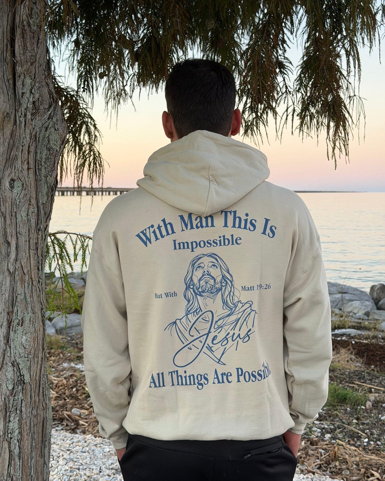 A man viewed from behind wearing a cream-colored hoodie with a blue graphic print on the back, standing outdoors near a tree and water at sunset.
