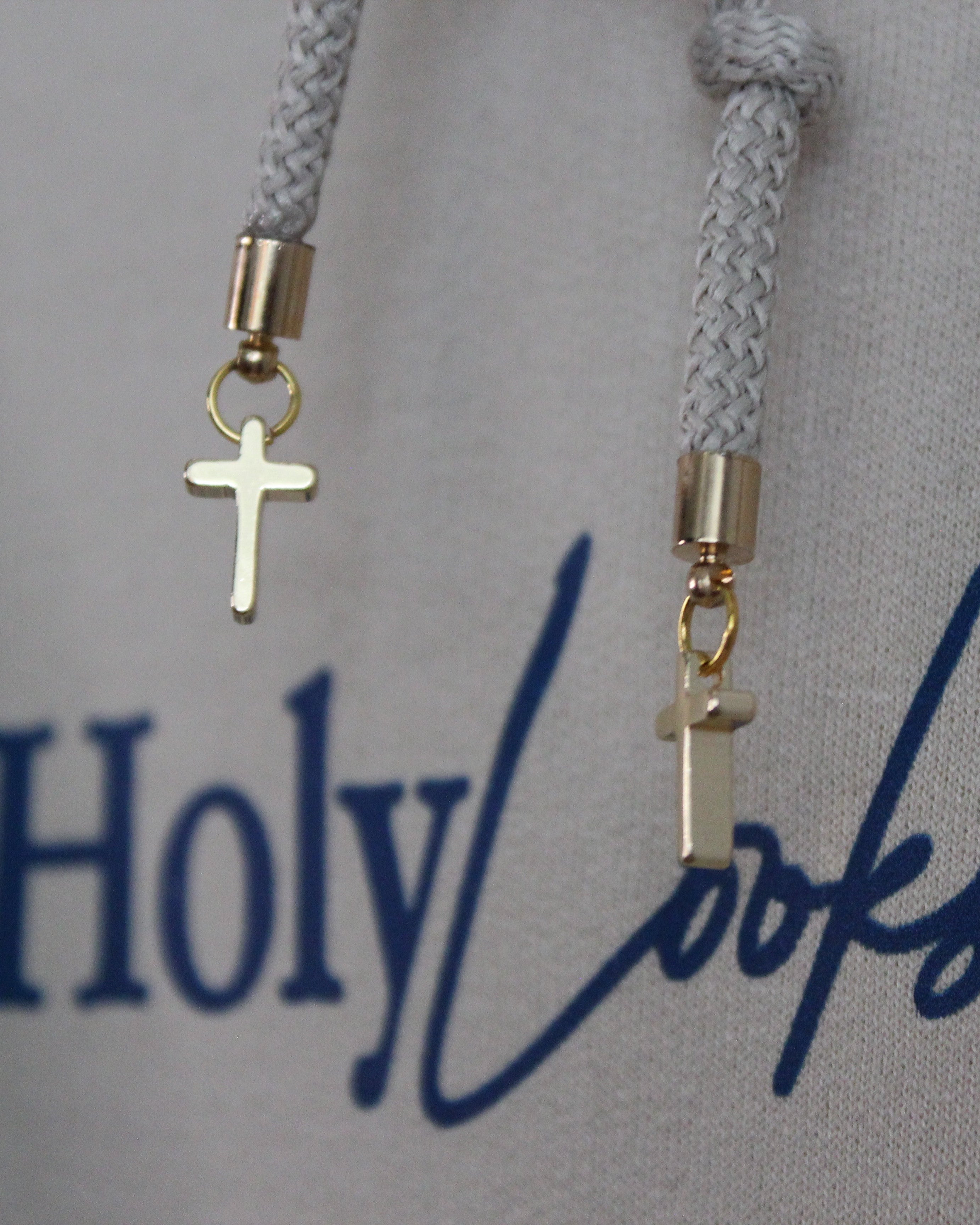 Close-up of a light gray hoodie with a dark blue 'Holy Looks' graphic print and white drawstrings adorned with small gold cross charms.
