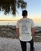 A man stands on a pebble beach facing the ocean at sunset, wearing a cream-colored t-shirt with a blue graphic and text on the back that reads 'With Man This Is Impossible All Things Are Possible'.