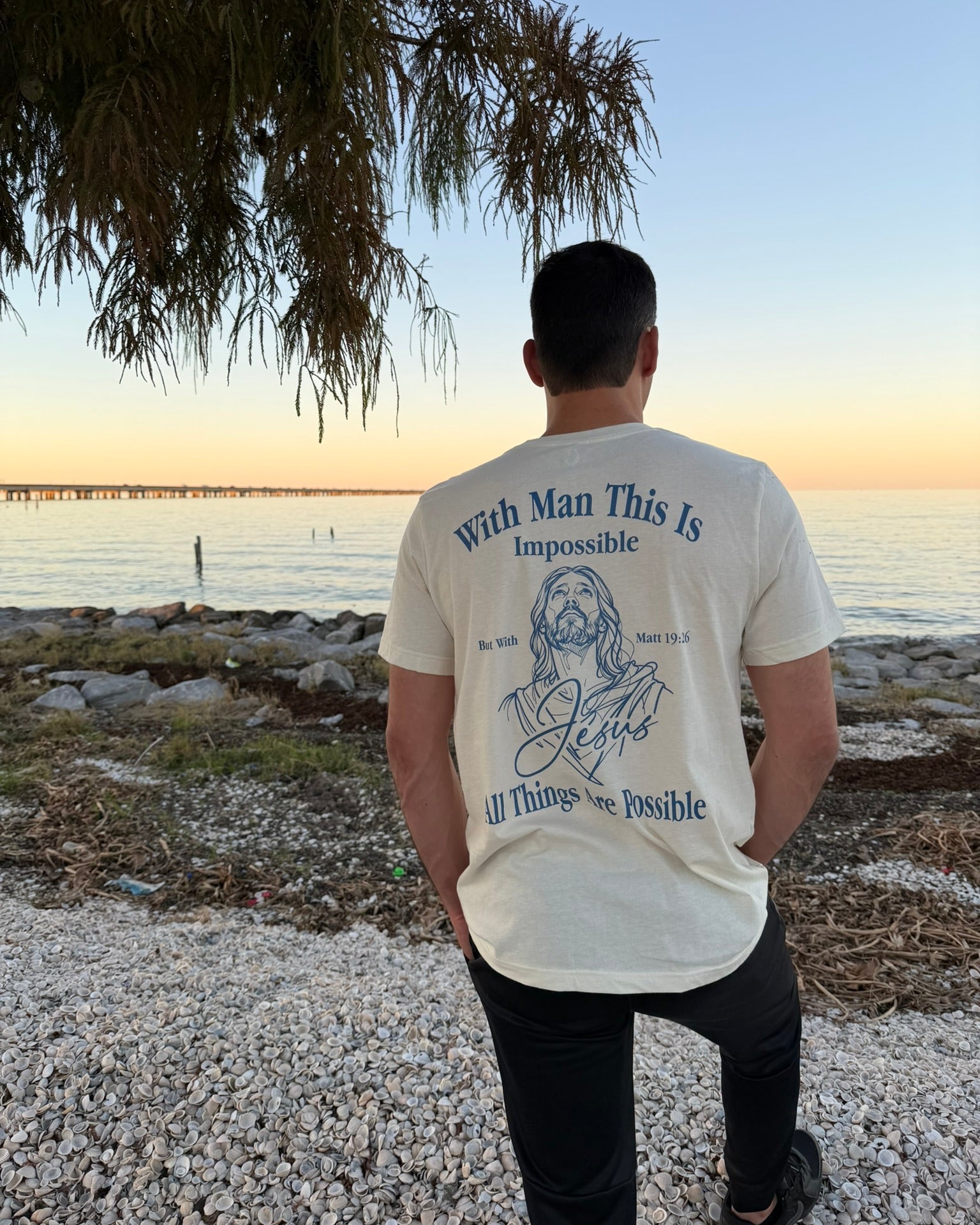 A man stands on a pebble beach facing the ocean at sunset, wearing a cream-colored t-shirt with a blue graphic and text on the back that reads 'With Man This Is Impossible All Things Are Possible'.