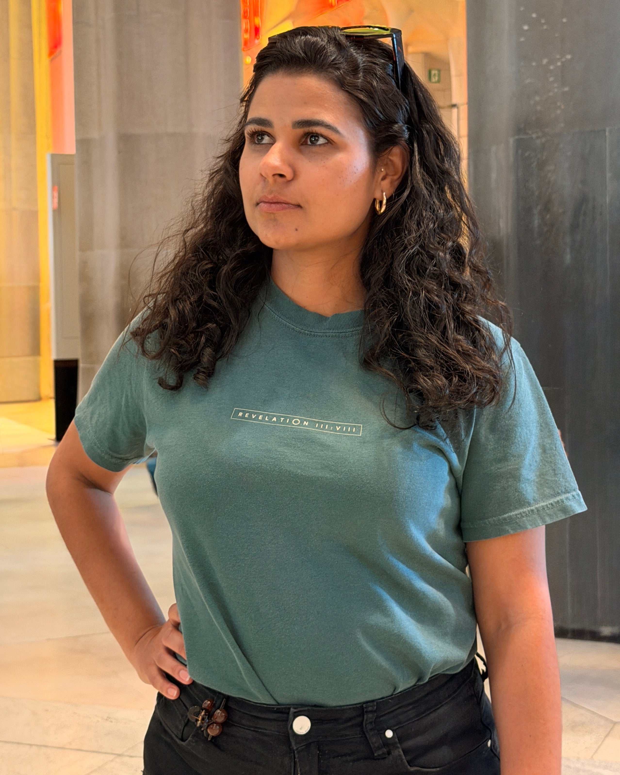 A woman with long, curly dark hair wearing a muted teal graphic t-shirt and black jeans, standing in a modern indoor setting with stone pillars and warm-toned lighting.