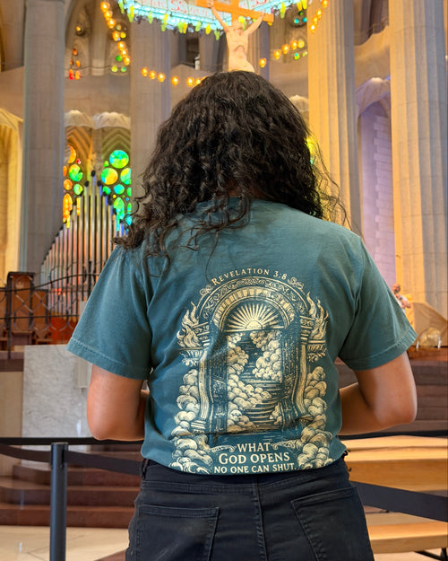 A person viewed from behind wearing a teal graphic T-shirt with a large cream-colored religious illustration and text on the back, standing inside a cathedral with stained glass windows and an organ in the background.