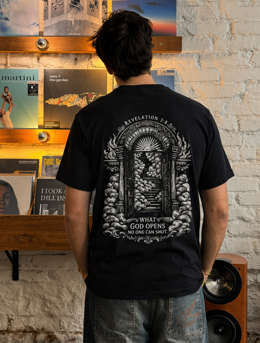 A person viewed from behind wearing a black graphic t-shirt and wide-leg jeans, standing in a record store with vinyl records displayed on shelves against a white brick wall.