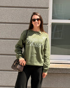A woman wearing an olive green crewneck sweatshirt with 'FOUND IN Christ' printed in white across the front, paired with black pants and sunglasses, standing outdoors against a gray stone wall.
