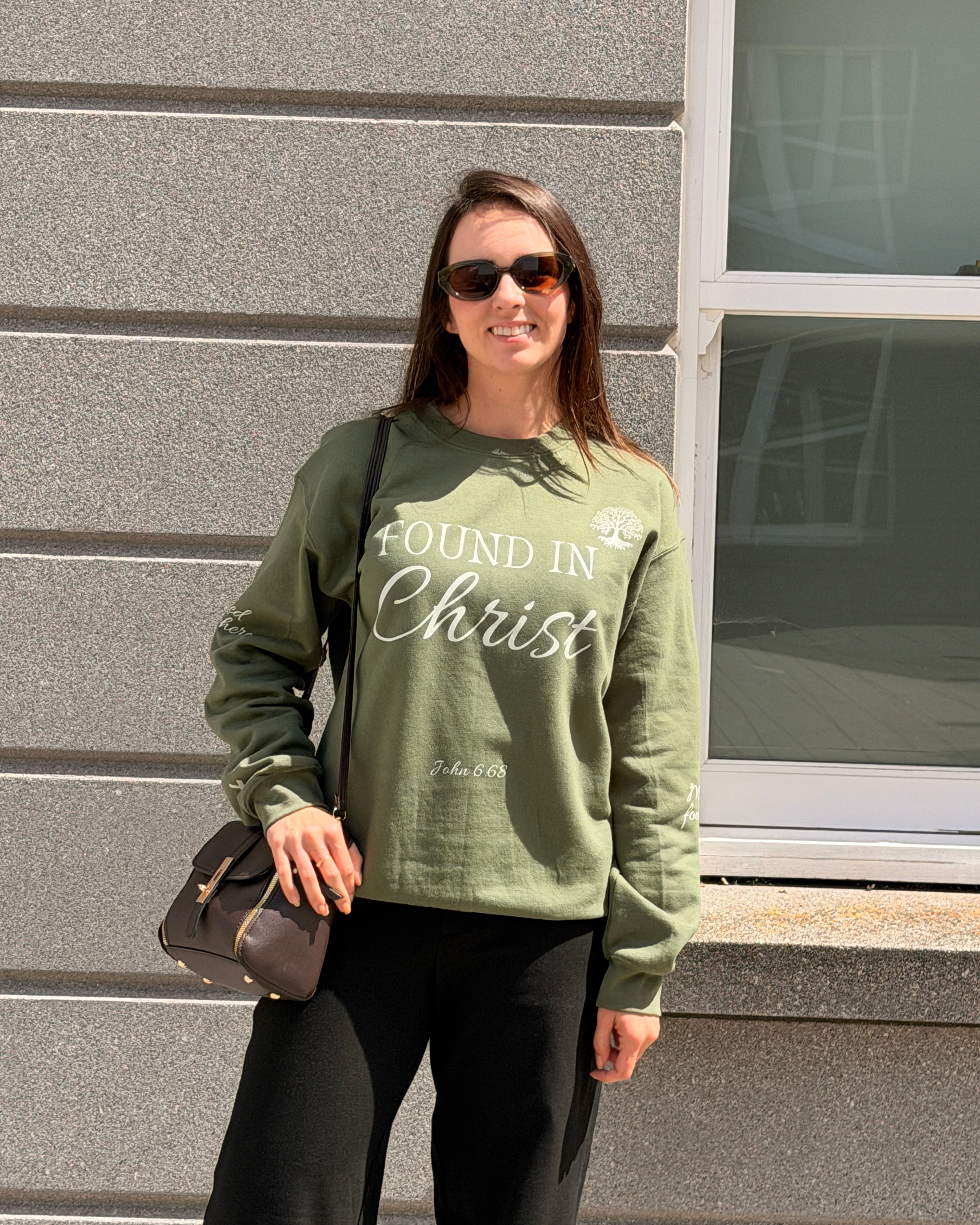 A woman wearing an olive green crewneck sweatshirt with 'FOUND IN Christ' printed in white across the front, paired with black pants and sunglasses, standing outdoors against a gray stone wall.