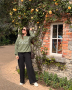 A woman wearing an olive green sweatshirt with 'I Found My Christ' printed on the front, black wide-leg pants, and white sneakers stands on a brick path beside a stone wall covered in yellow roses.