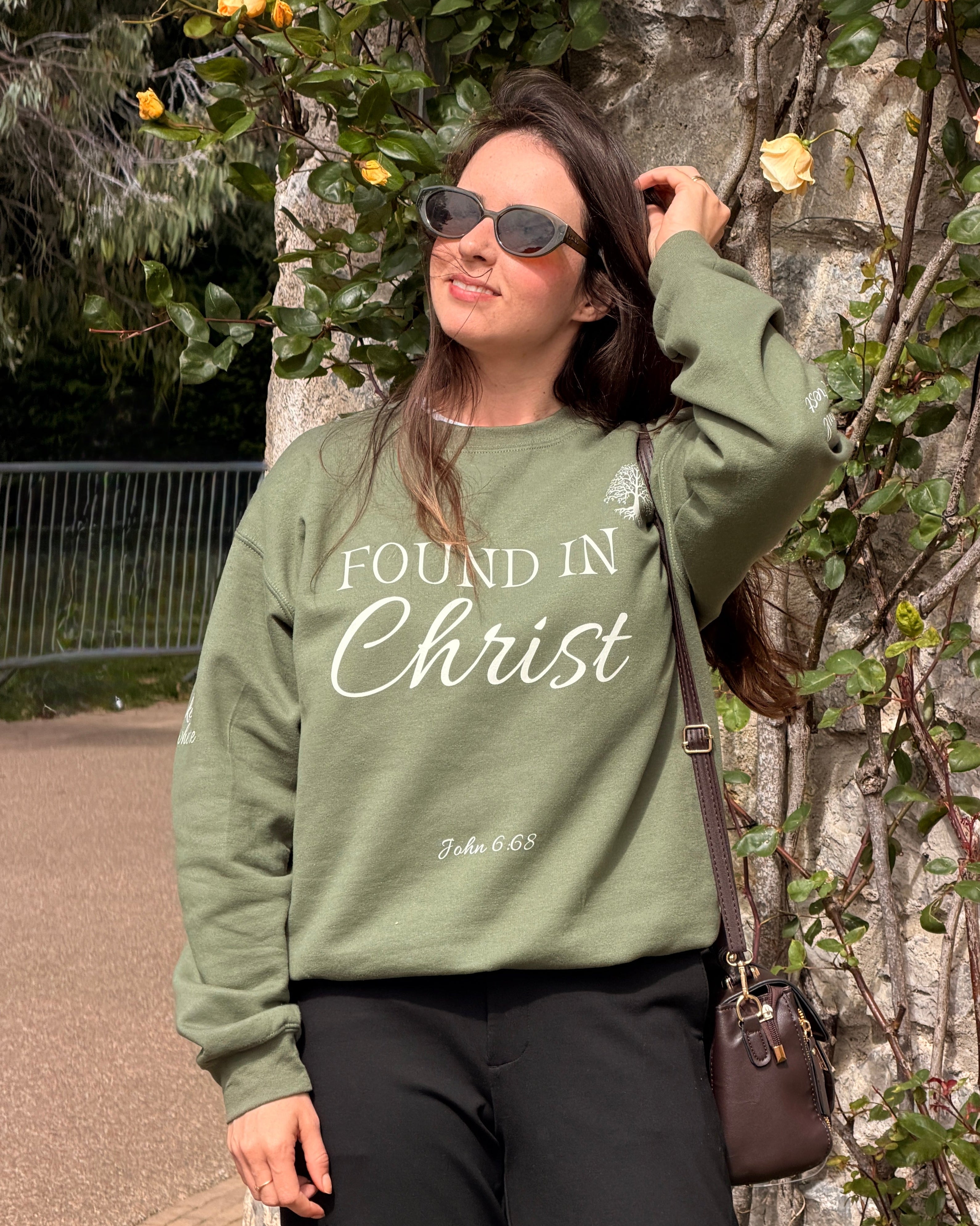 A woman wearing an olive green crewneck sweatshirt with 'FOUND IN Christ' printed in white script, standing outdoors against a brick wall and climbing roses.
