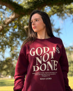A woman wearing a burgundy sweatshirt with the text 'GOD IS NOT DONE' in white, standing outdoors under a large tree in a park.