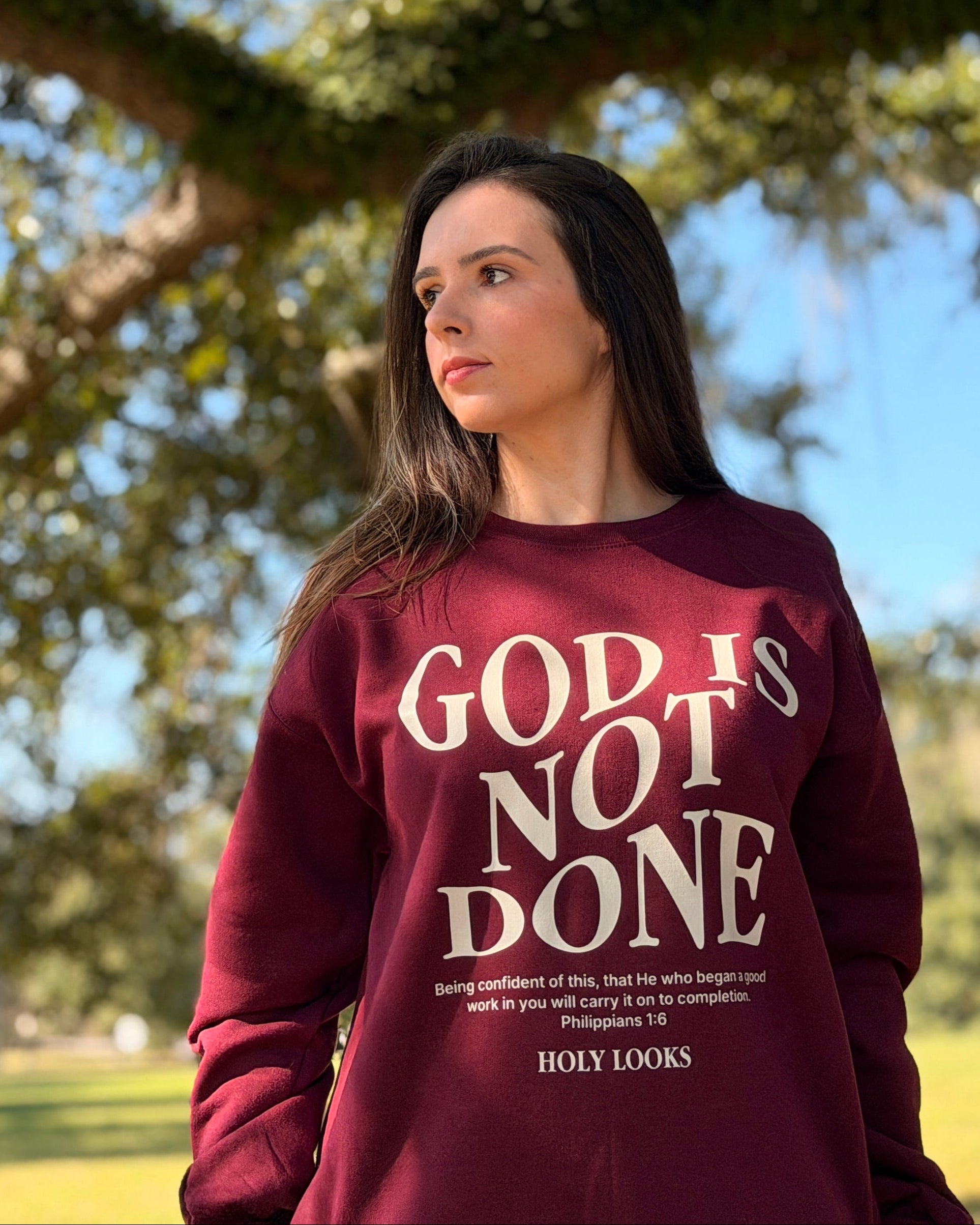 A woman wearing a burgundy sweatshirt with the text 'GOD IS NOT DONE' in white, standing outdoors under a large tree in a park.