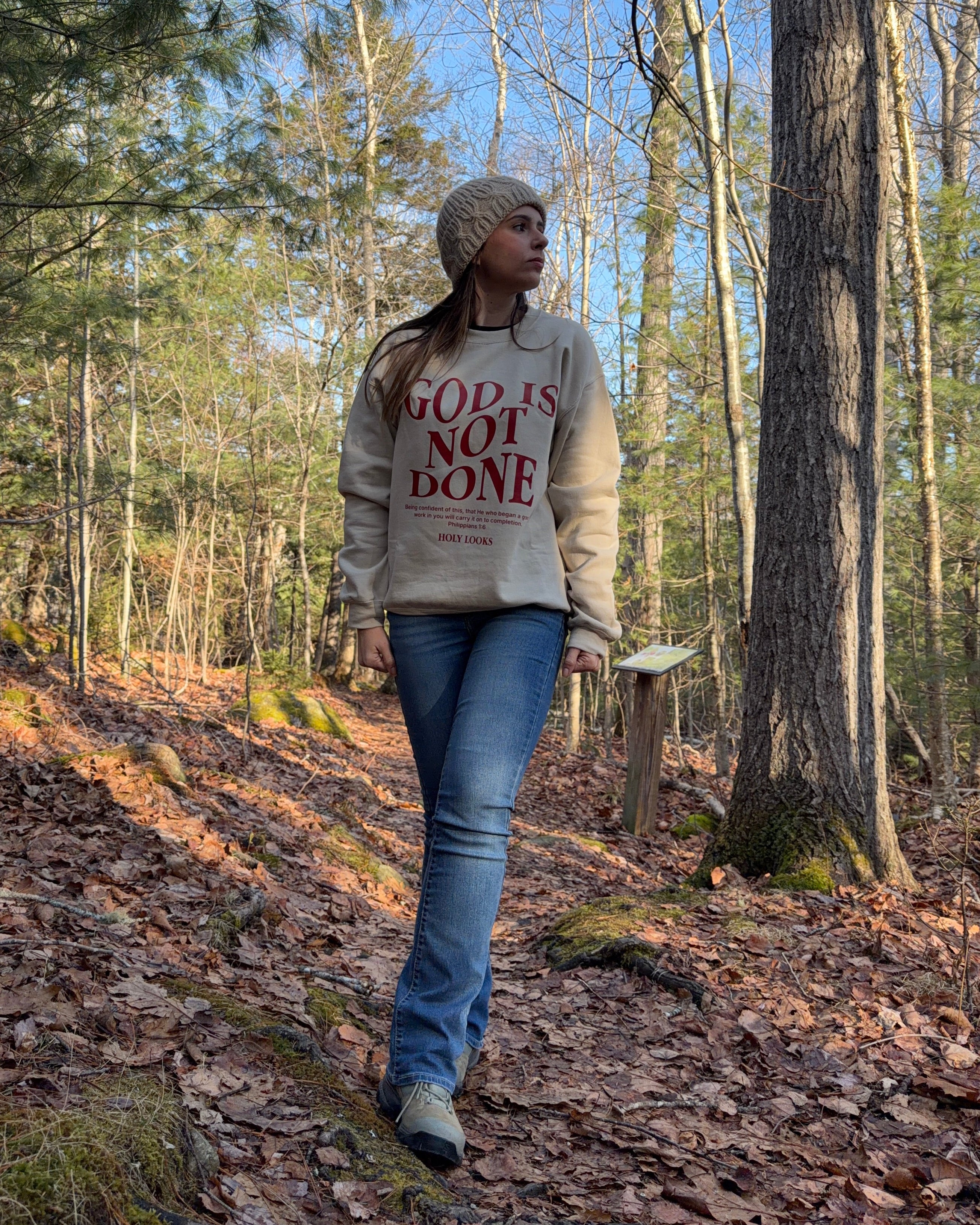 A woman wearing a beige graphic sweatshirt with the text 'GOD IS NOT DONE' in red, blue jeans, and a knit beanie walks along a leaf-covered forest path.