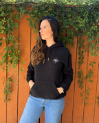 A woman wearing a black pullover hoodie with a white embroidered logo on the chest, paired with light blue jeans, standing in front of an orange-brown wooden fence with green ivy.