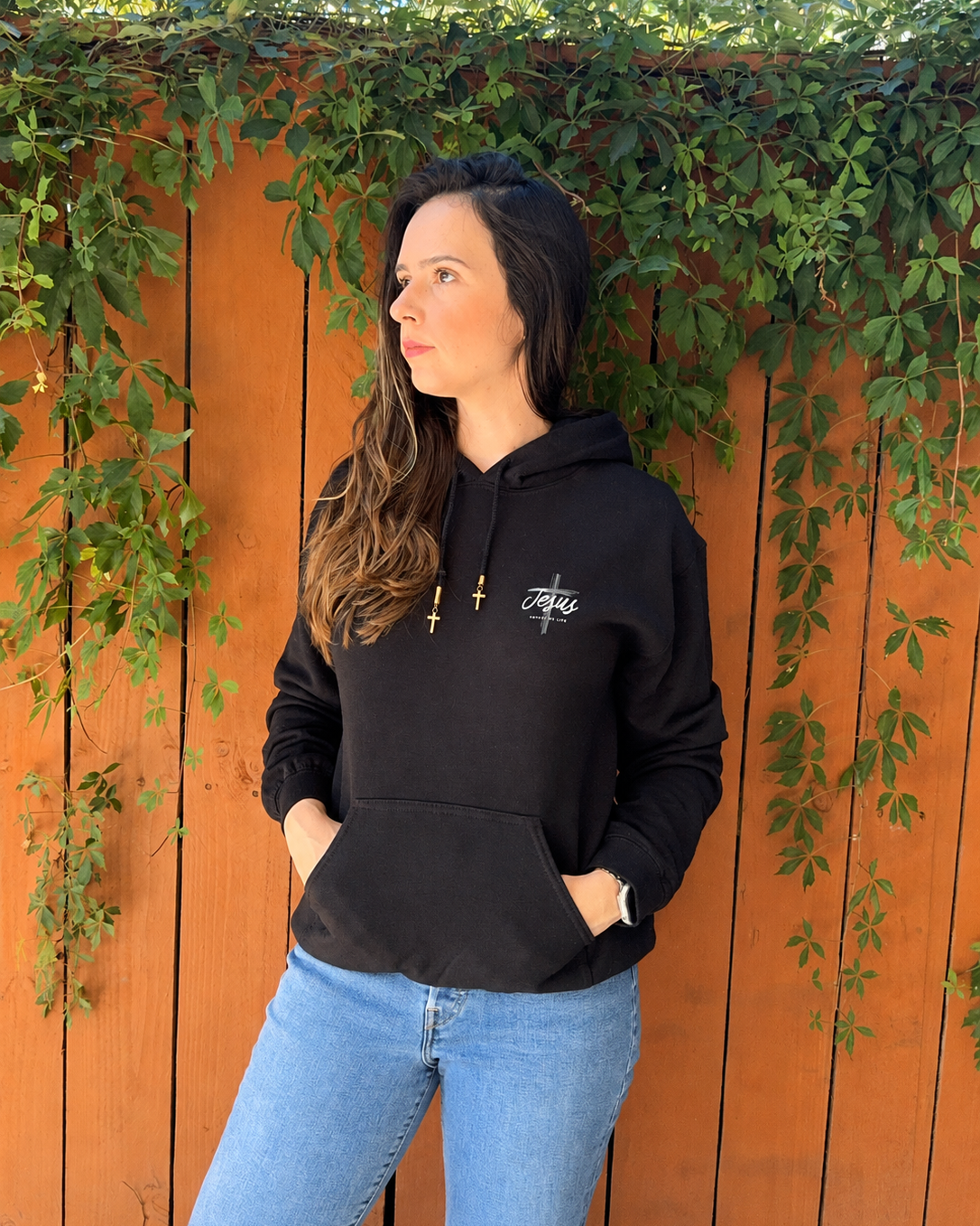 A woman wearing a black pullover hoodie with a white embroidered logo on the chest, paired with light blue jeans, standing in front of an orange-brown wooden fence with green ivy.