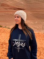 A woman wearing a cream knit beanie and a navy blue sweatshirt with a white graphic reading 'Jesus saves my life' stands in a desert landscape with red rock formations and orange sand under an overcast sky.