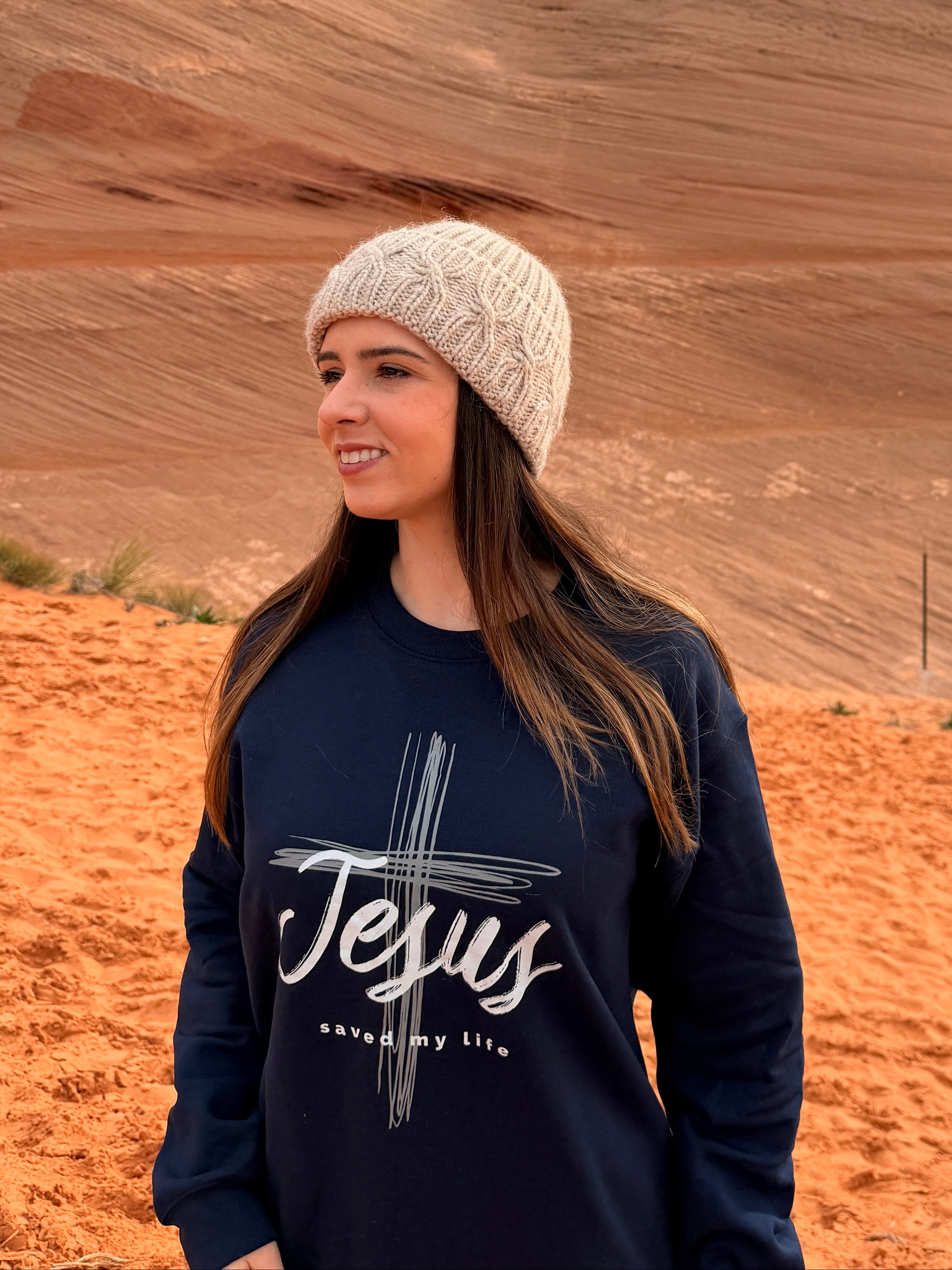 A woman wearing a cream knit beanie and a navy blue sweatshirt with a white graphic reading 'Jesus saves my life' stands in a desert landscape with red rock formations and orange sand under an overcast sky.