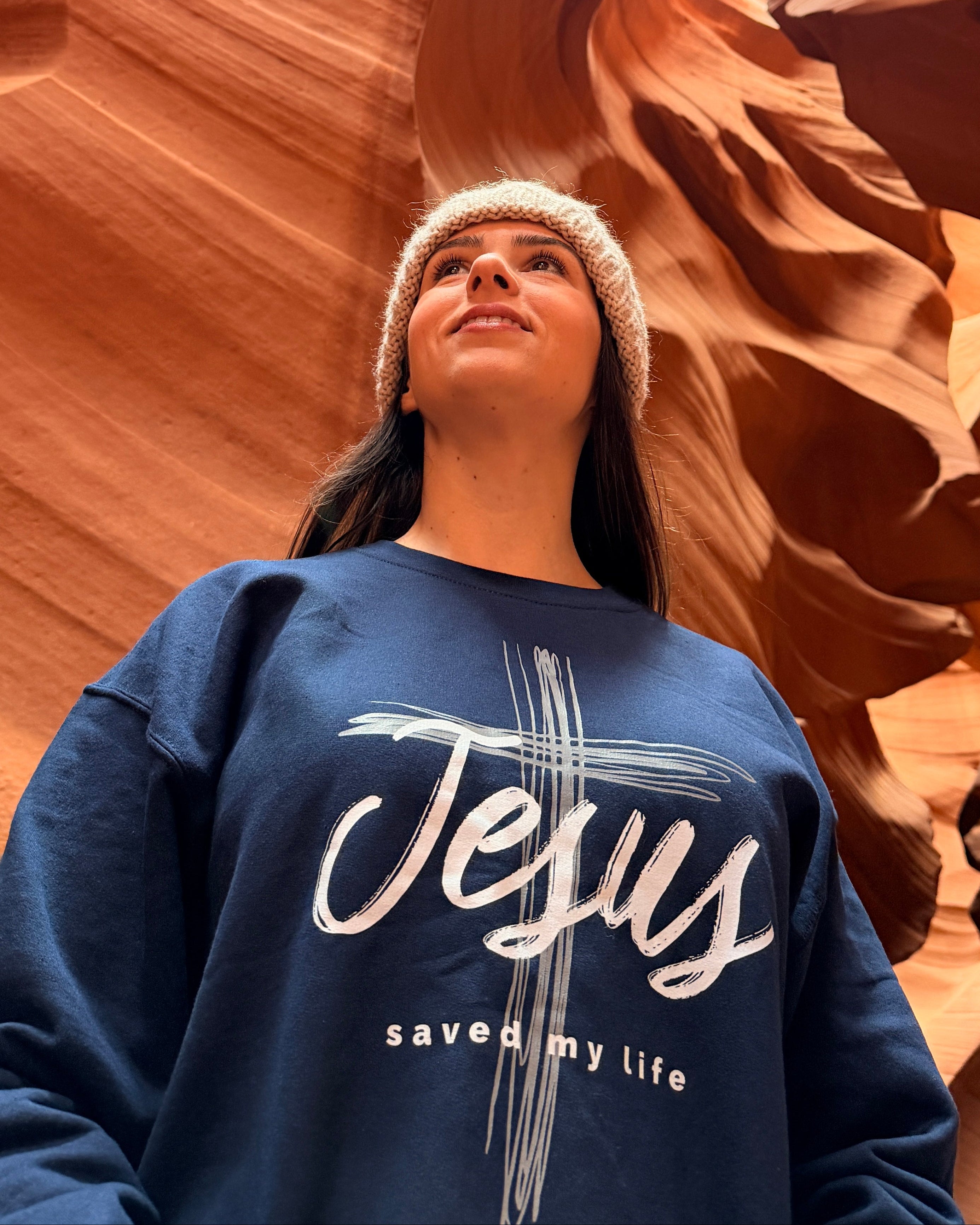 A woman wearing a navy blue sweatshirt with 'Jesus saved my life' printed on the front, standing inside a slot canyon with orange sandstone walls.