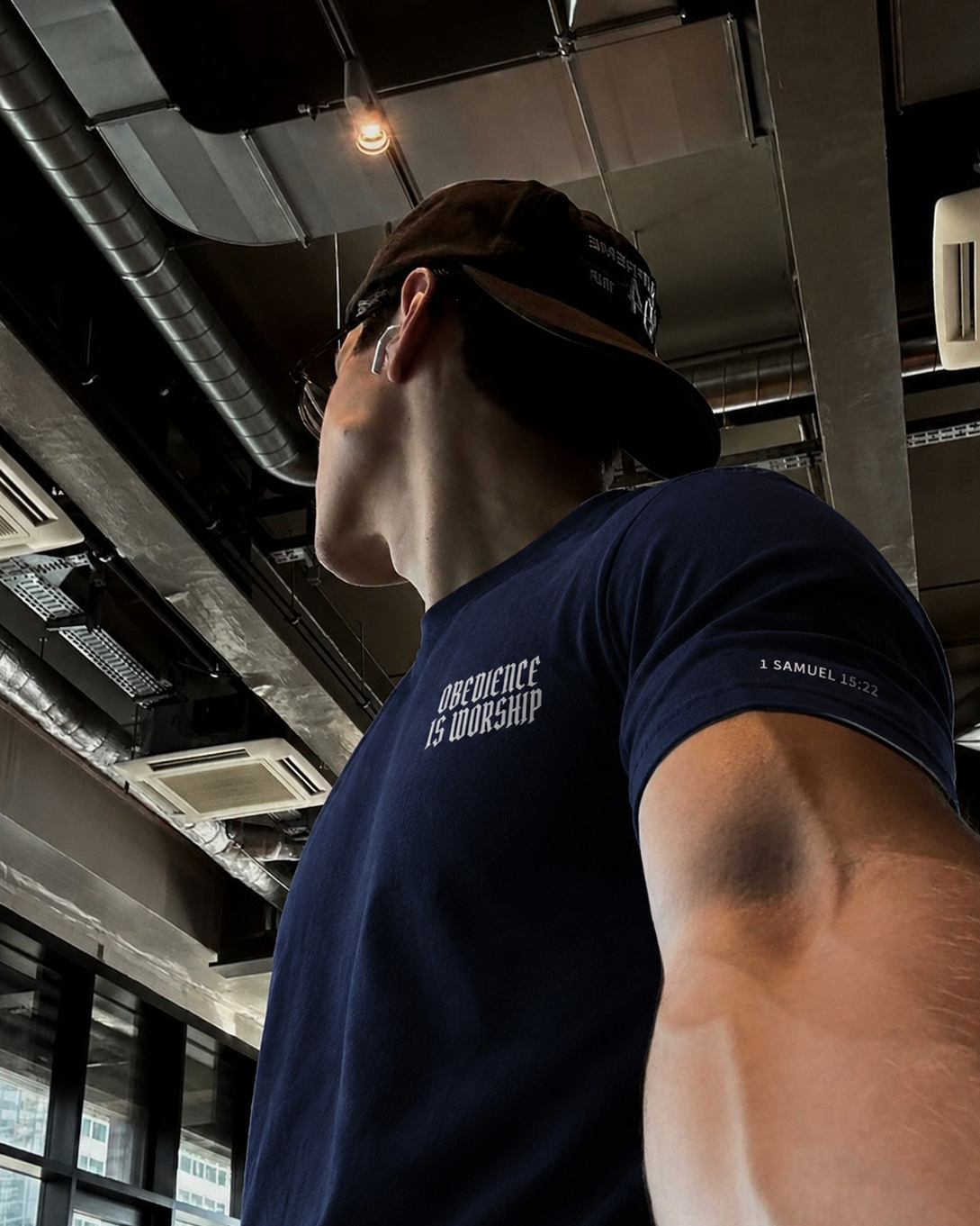A man wearing a navy blue t-shirt with the text 'OBEDIENCE IS WORSHIP' on the chest and a brown baseball cap, photographed from a low angle in an industrial gym setting.