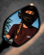 A person wearing a black full-face helmet and a dark burgundy t-shirt with white text, seen in a motorcycle side mirror. The background is a close-up of the motorcycle's dark frame.