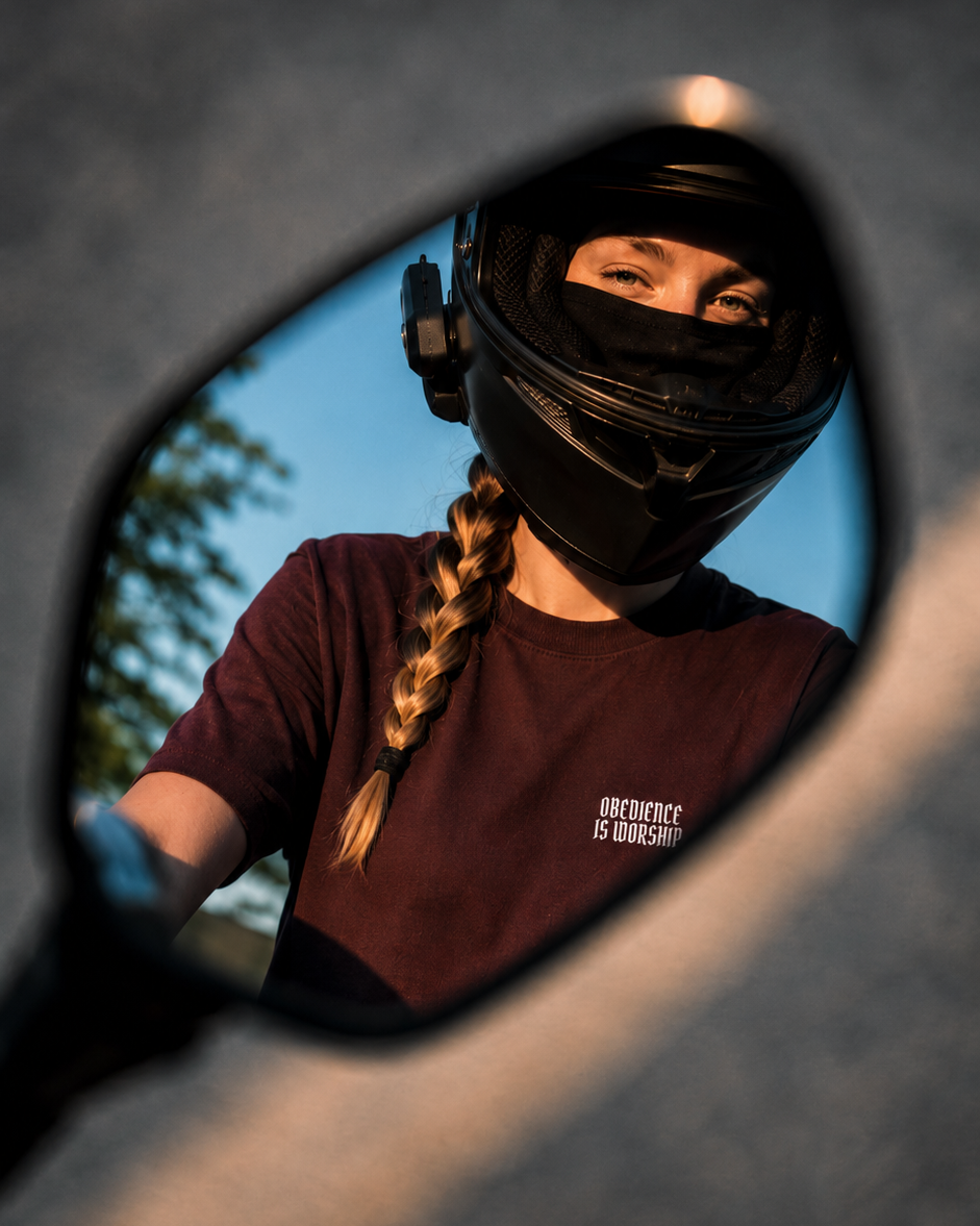 A person wearing a black full-face helmet and a dark burgundy t-shirt with white text, seen in a motorcycle side mirror. The background is a close-up of the motorcycle's dark frame.
