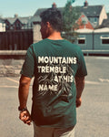 A man viewed from behind wearing a dark forest green t-shirt with a large white graphic print on the back that reads 'MOUNTAINS TREMBLE AT HIS NAME' with a mountain illustration and 'Psalm 85'.