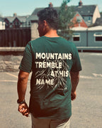 A man viewed from behind wearing a dark forest green t-shirt with a large white graphic print on the back that reads 'MOUNTAINS TREMBLE AT HIS NAME' with a mountain illustration and 'Psalm 85'.