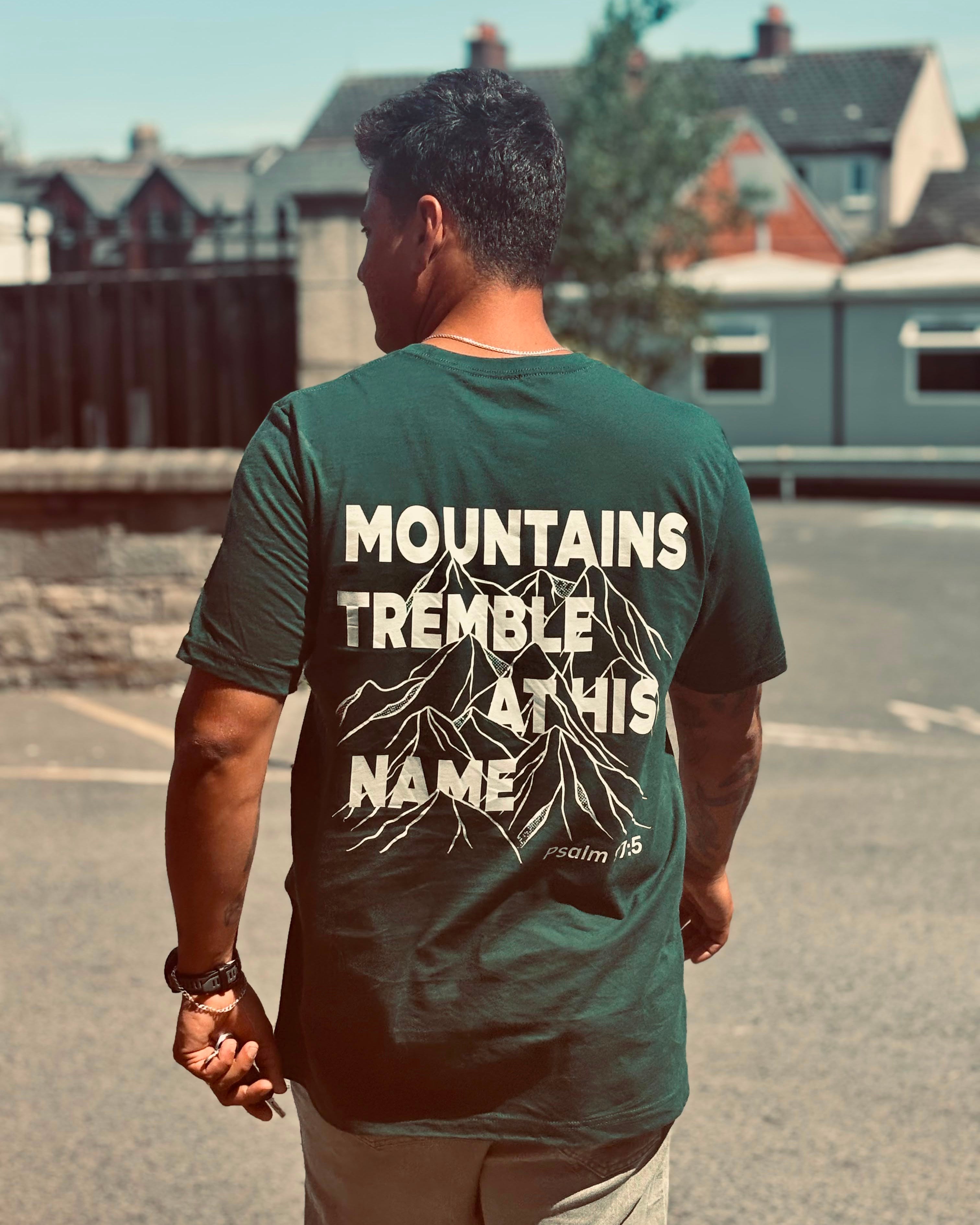 A man viewed from behind wearing a dark forest green t-shirt with a large white graphic print on the back that reads 'MOUNTAINS TREMBLE AT HIS NAME' with a mountain illustration and 'Psalm 85'.