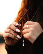A close-up lifestyle shot of a person holding a black cord necklace with a small gold cross pendant. The person has long red hair, white manicured nails, and is wearing a gold ring on one finger.