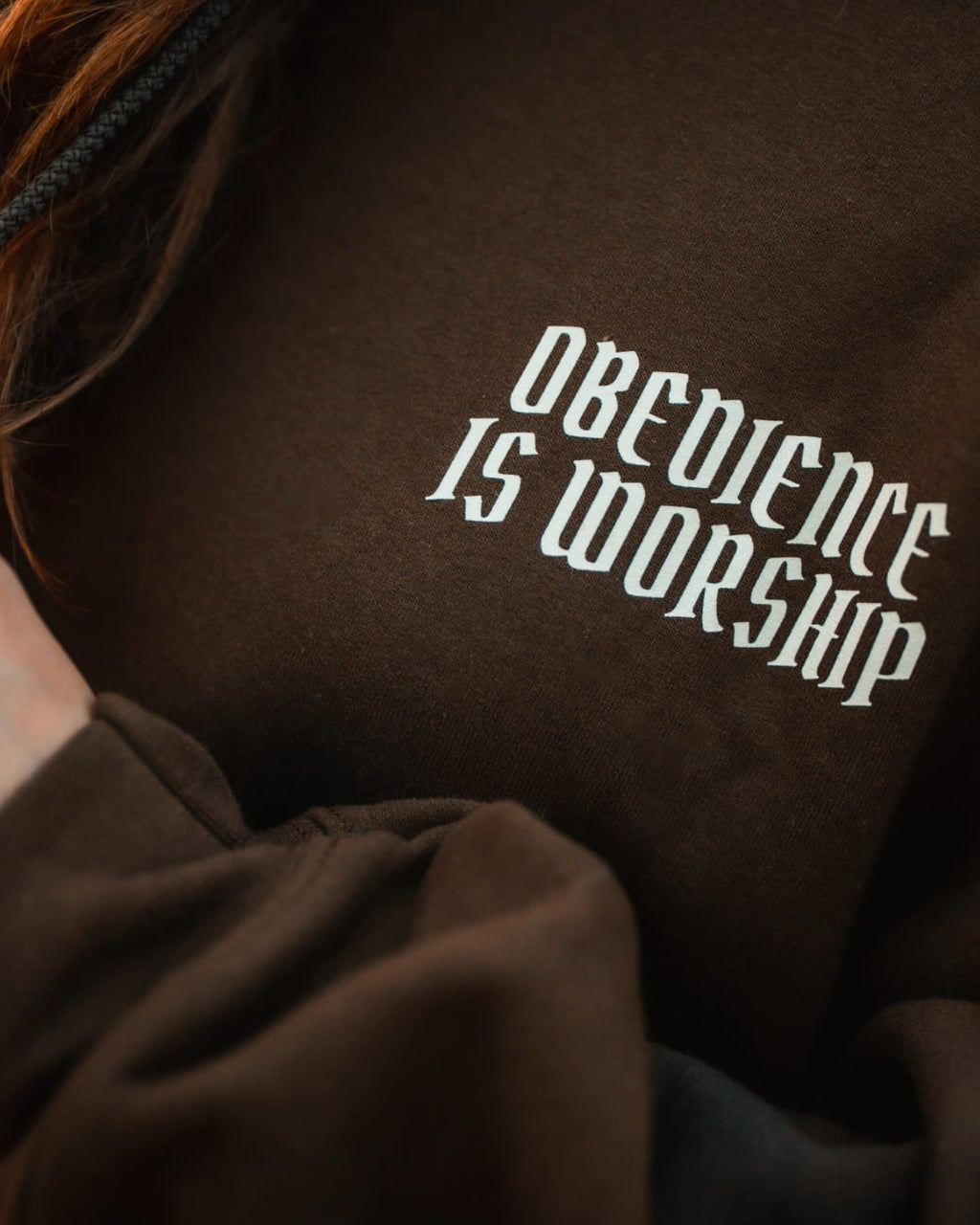 Close-up of a person wearing a dark brown hoodie with white gothic-style text reading 'Obedience is Worship' on the chest.