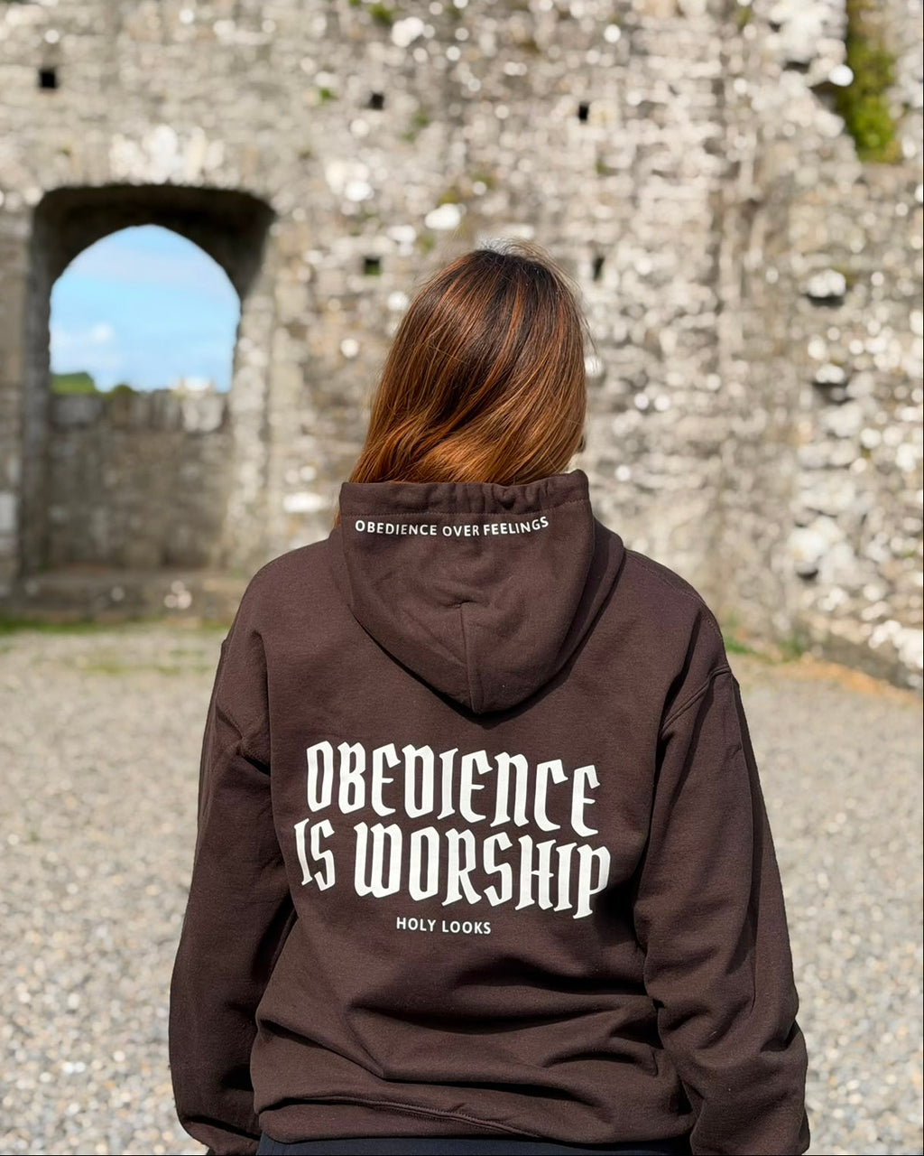 A person viewed from behind wearing a dark brown hoodie with 'Obedience is Worship' printed in white on the back, standing in front of a stone castle ruin.
