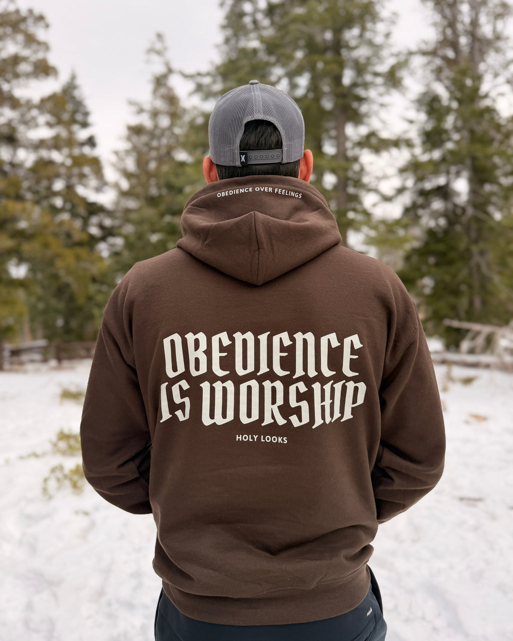 A man viewed from behind wearing a brown hoodie with 'OBEIDENCE IS WORSHIP' printed in white on the back, standing in a snowy outdoor setting with trees in the background.