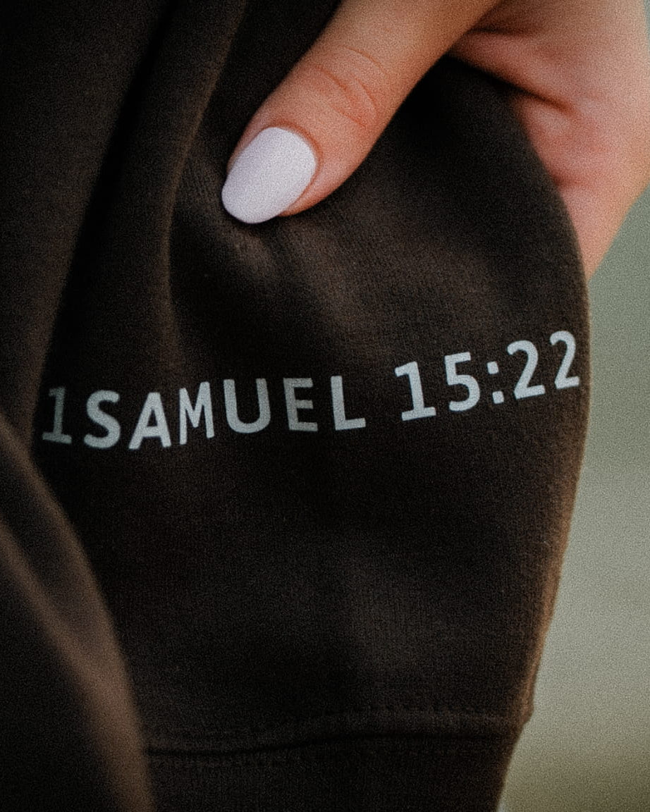Close-up of a person wearing a dark brown hoodie, showing the sleeve with the text '1 SAMUEL 15:22' printed in white. A hand with light pink nail polish is holding the fabric.