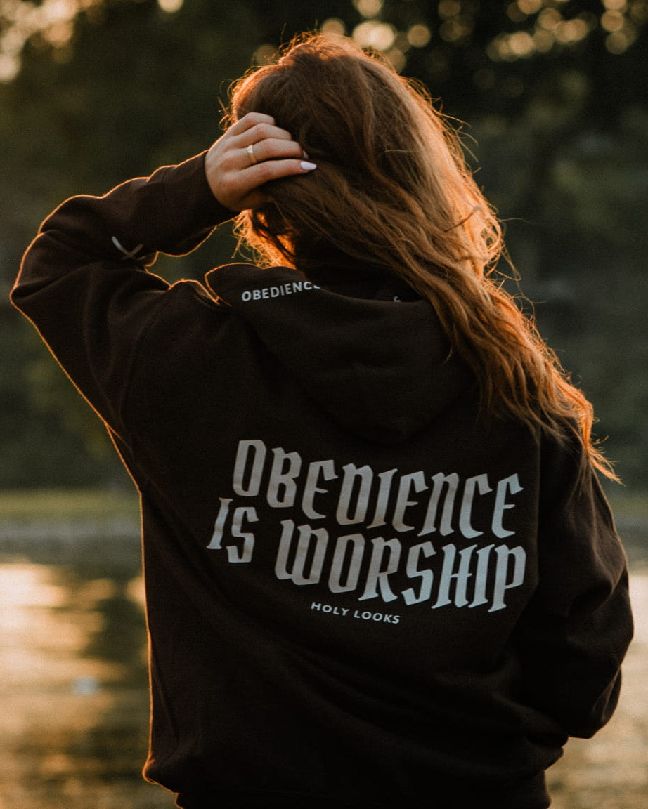 A woman with long brown hair stands with her back to the camera by a body of water at sunset, wearing a black hoodie with 'OBEDIENCE IS WORSHIP' printed in white on the back.