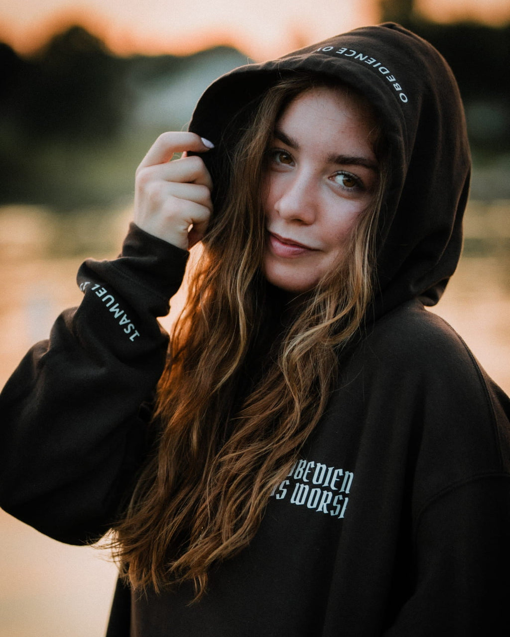 A woman wearing a black hoodie with white text on the hood and sleeve, photographed outdoors at sunset with a blurred background.