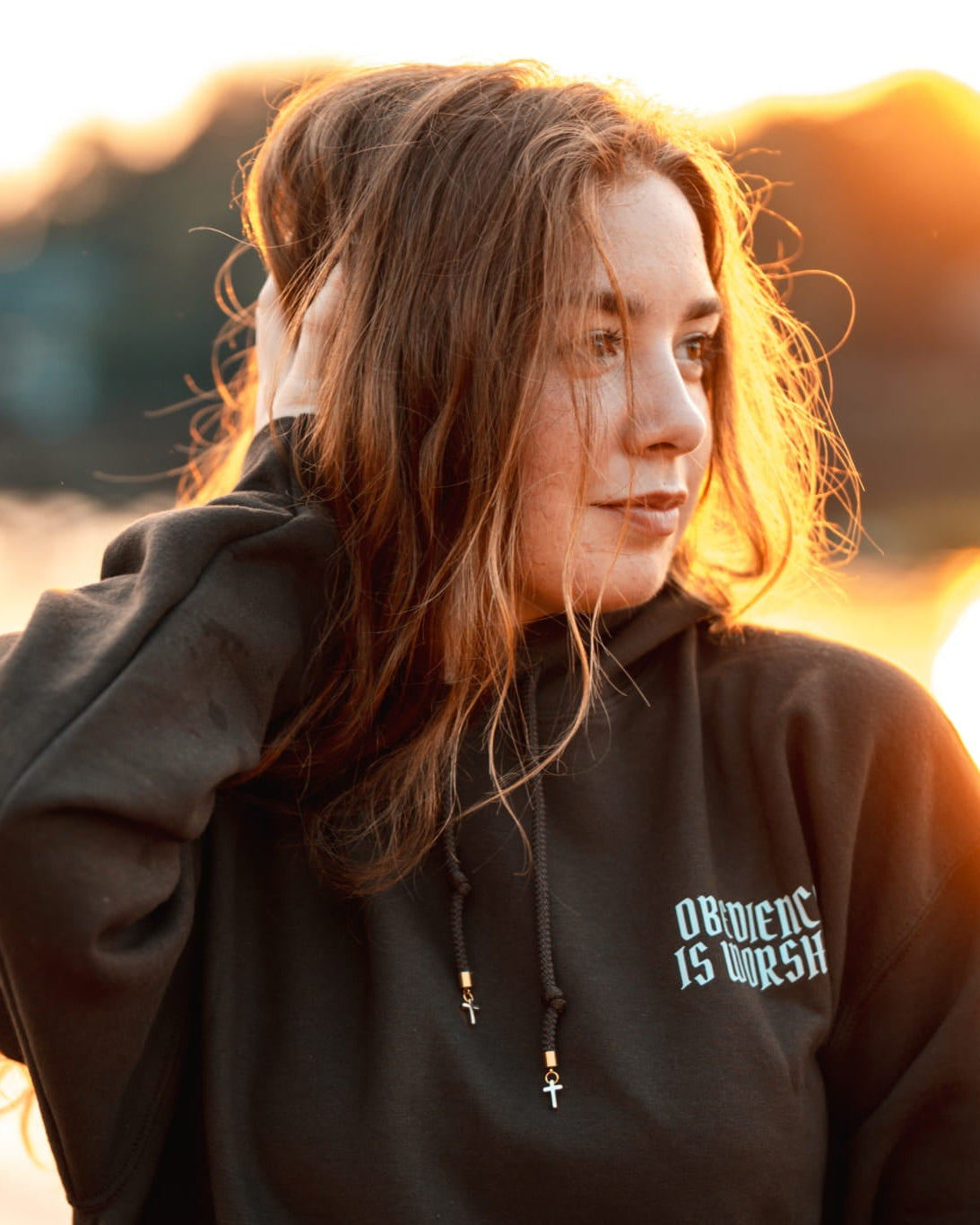 A woman with long light brown hair stands outdoors at sunset wearing a black hoodie with white gothic text on the left chest that reads 'OBSCURE IS UPSIT'. The background shows a warm, glowing body of water with blurred trees and hills.