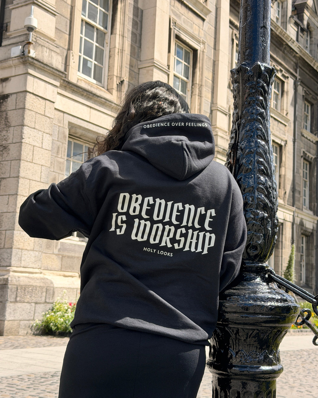 A person viewed from behind wearing a black hoodie with 'OBEDIENCE IS WORSHIP' printed in white across the back, standing outdoors next to a black lamppost in front of a stone building.
