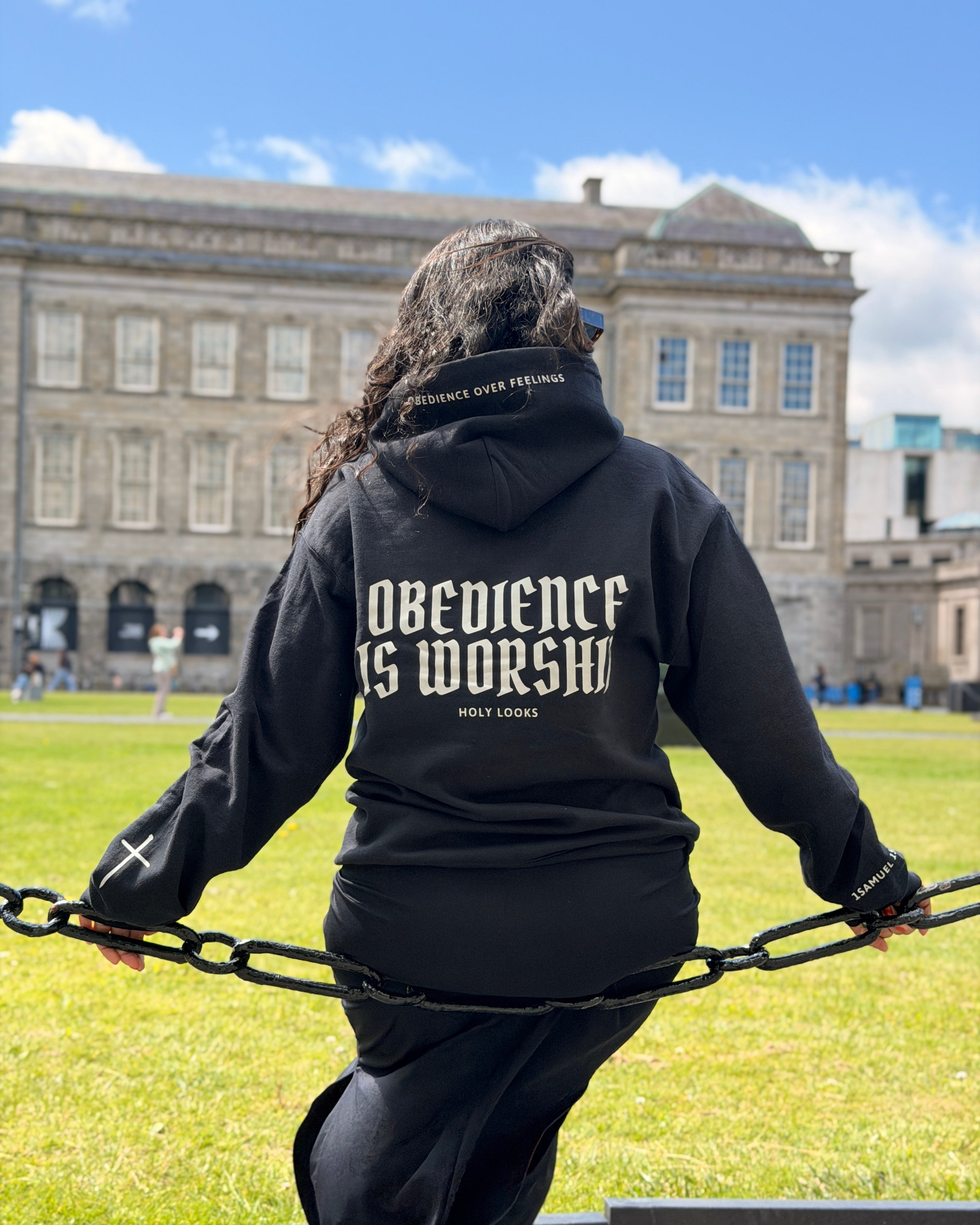 A person viewed from behind wearing a black hoodie with 'OBEDIENCE IS WORSHIP' printed in white across the back, standing outdoors on a grassy lawn in front of a large stone building under a blue sky.