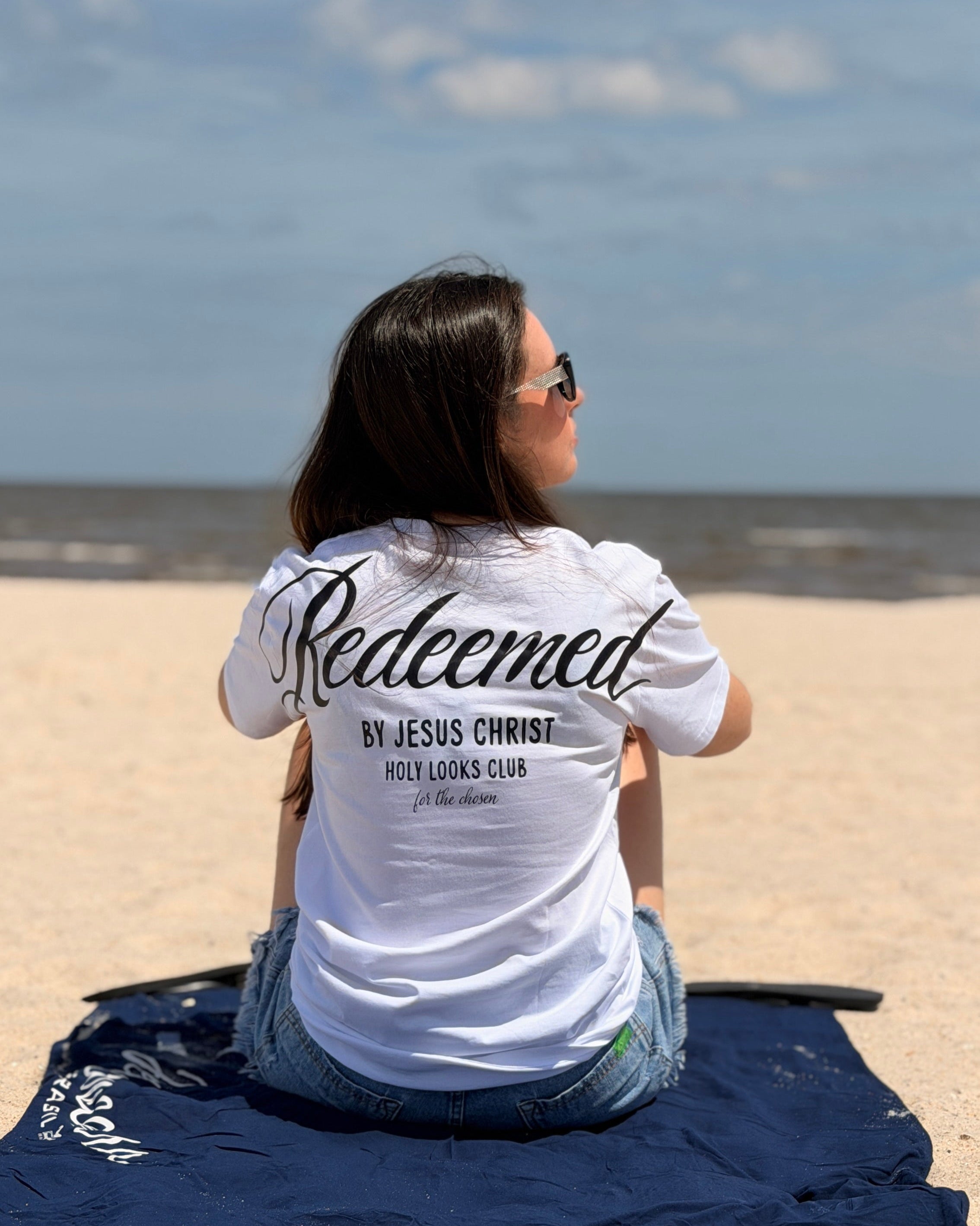 A woman sits on a navy blue towel on a sandy beach, viewed from behind. She is wearing a white t-shirt with 'Redeemed BY JESUS CHRIST WALK LOOKS CLUB' printed in black script on the back.