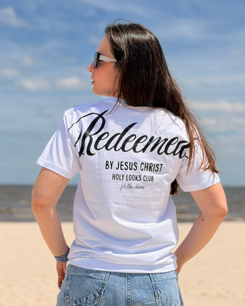 A woman standing on a sandy beach with her back to the camera, wearing a white t-shirt with black text that reads 'Redeemer BY JESUS CHRIST HOLY LOOKS CLUB' and light blue jeans.