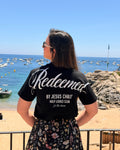 A woman stands on a beach facing the ocean, wearing a black t-shirt with 'Redeemed BY JESUS CHRIST HOLY LOOKS CLUB ft. the chums' printed in white on the back, paired with a black floral skirt.