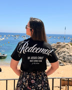 A woman stands on a beach facing the ocean, wearing a black t-shirt with 'Redeemed BY JESUS CHRIST HOLY LOOKS CLUB ft. the chums' printed in white on the back, paired with a black floral skirt.