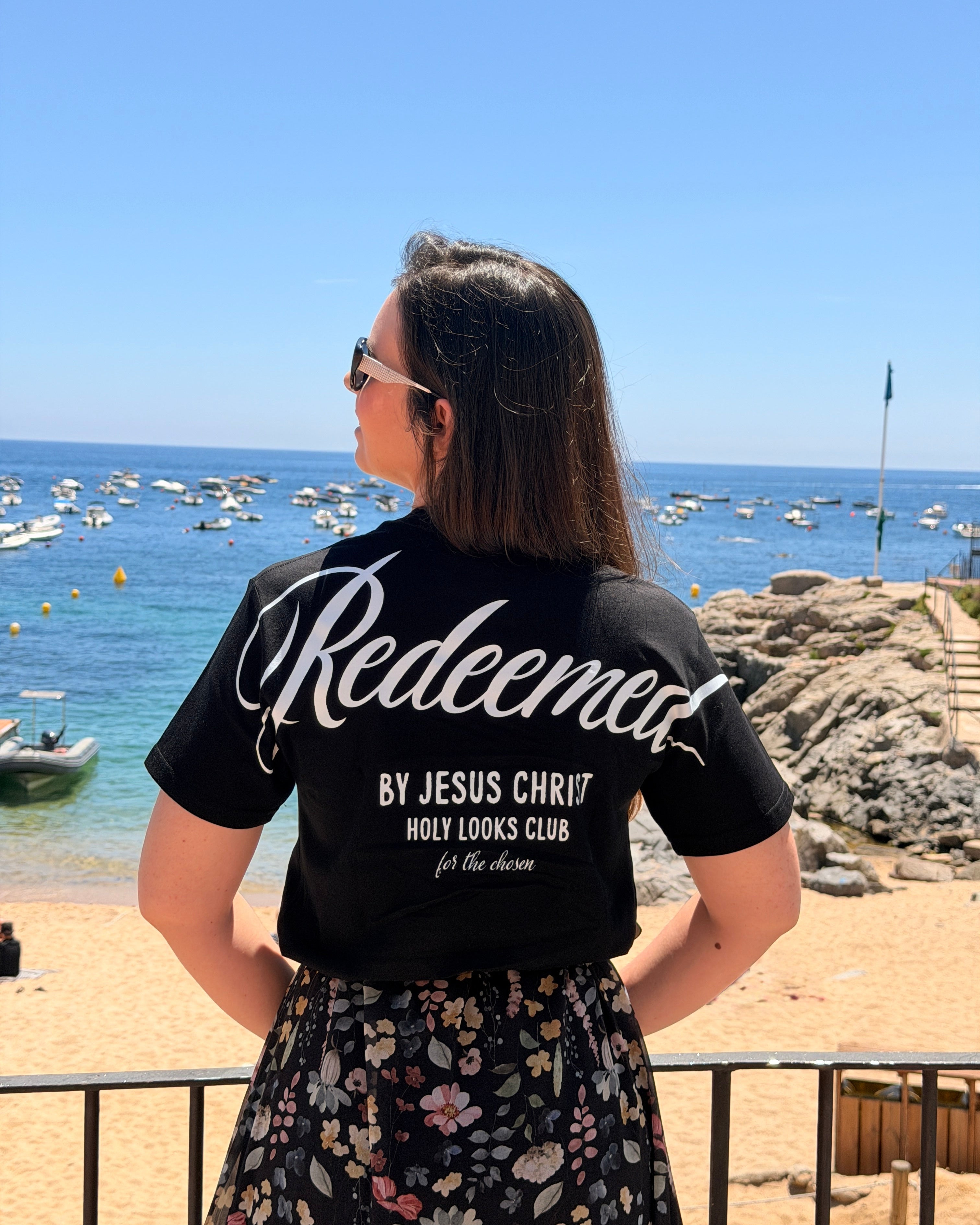 A woman stands on a beach facing the ocean, wearing a black t-shirt with 'Redeemed BY JESUS CHRIST HOLY LOOKS CLUB ft. the chums' printed in white on the back, paired with a black floral skirt.
