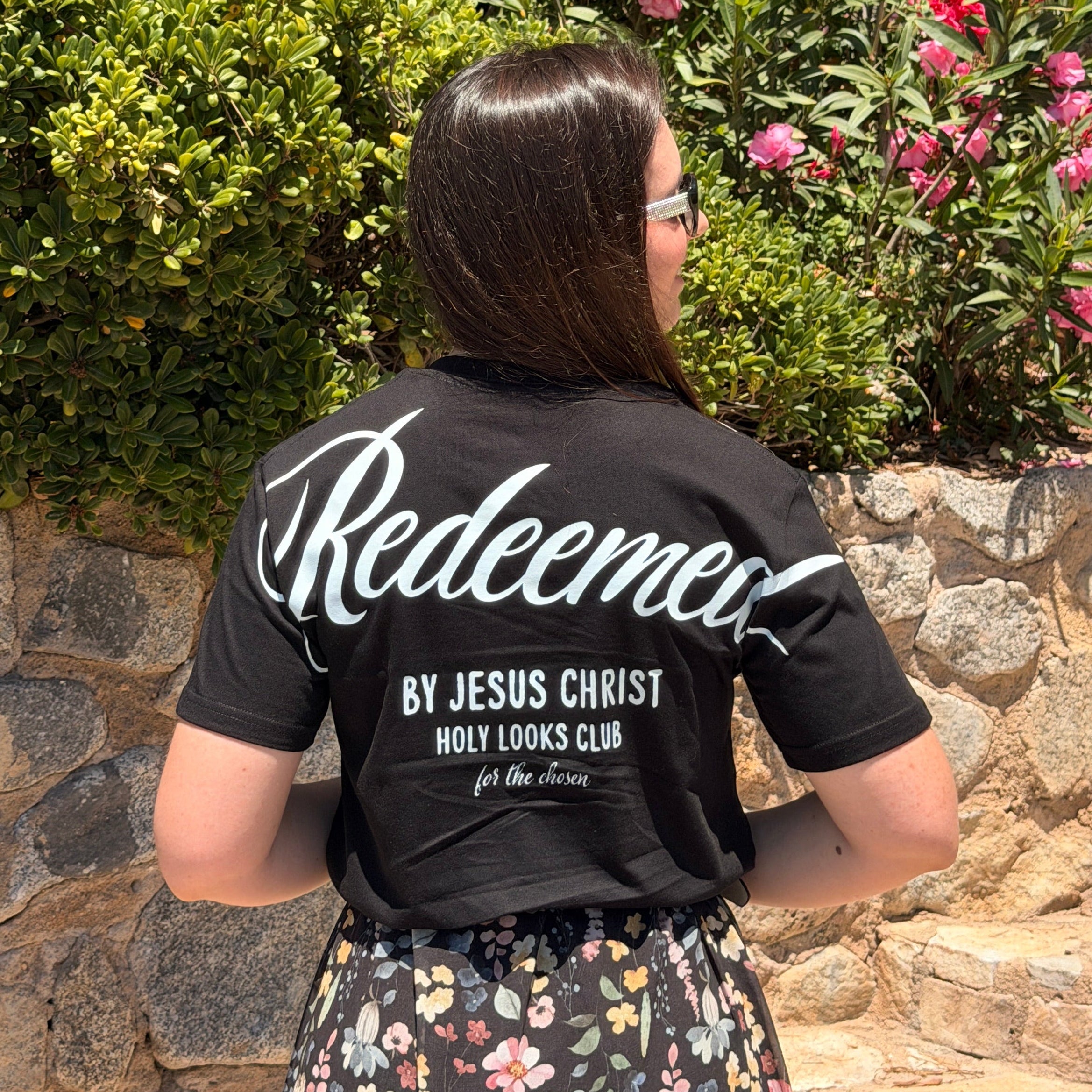 A woman viewed from behind wearing a black graphic t-shirt with 'Redeemer BY JESUS CHRIST HOLY LOOKS CLUB' printed in white on the back, standing outdoors in front of a stone wall and green bushes with pink flowers.